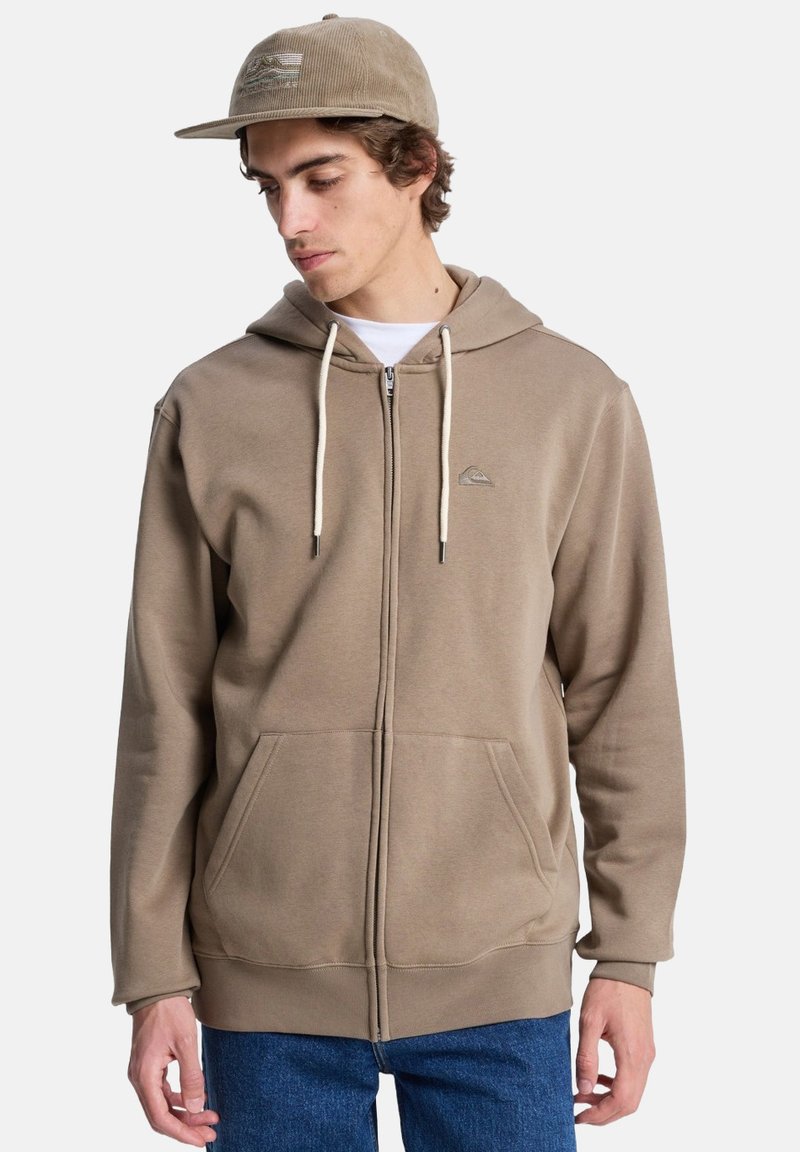Bruine zip-up hoodie met een trekkoordkap, twee voorkwapen en een subtiele logo. Gecombineerd met een lichtbeige pet met een gestreept ontwerp.