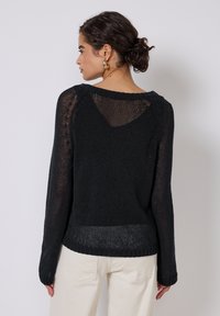 Maglione nero a maglia caratterizzato da una vestibilità comoda, maniche lunghe e dettagli con schiena scoperta. La texture è leggera e leggermente trasparente. Abbinato a pantaloni chiari.