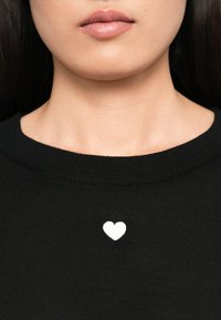 Mustasilmuke, jossa on pieni valkoinen sydänkuvio rintakehällä. Kaula-aukko on pyöreä, ja pinta vaikuttaa sileältä.