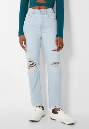 Lichtblauwe gescheurde jeans met grote scheuren op dijen en knieën, gecombineerd met witte sneakers en een teal kleurige top met lange mouwen die de buik laat zien.