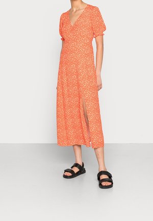 Femme portant une robe midi florale orange avec manches bouffantes et une fente avant, associée à des sandales noires à bout ouvert sur un sol gris.