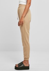 Beige joggebukser med snor i livet og smalnerende cuffs, parret med sorte platåsandaler, sett fra siden.