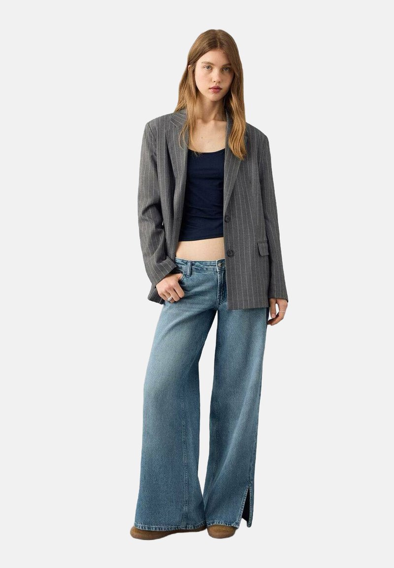 Gestreepte grijze blazer met een enkele knoop, gecombineerd met een marineblauwe croptop en lichtblauwe wijde jeans met zijsplitten. Bruine schoenen.