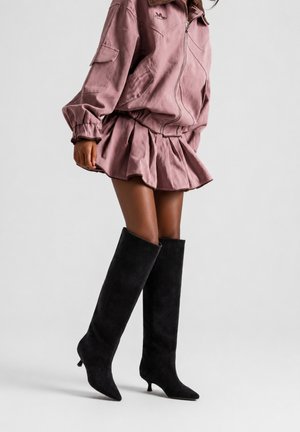 Femme portant une veste rose et une jupe avec de hautes bottes noires à talons, debout devant un fond clair uni.