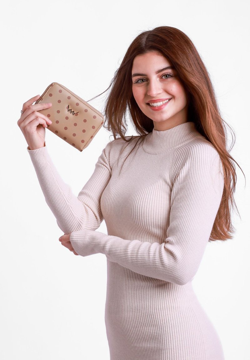 Beige wallet with brown polka dots and gold hardware logo, held in a hand; compact rectangular design with a textured surface.