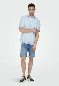 Hellblau gestreiftes Kurzarm-Hemd, kombiniert mit Jeansshorts mit aufgerollten Ärmeln. Schwarze Sneakers mit weißen Sohlen.