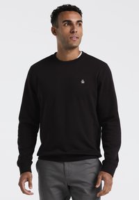 Zwarte sweatshirt met een ronde halslijn, ribgebreide cuffs en onderrand, voorzien van een klein wit logo op de linkerborst. Gecombineerd met lichtgrijze broek.
