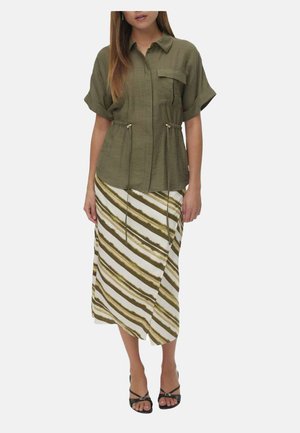 Femme portant une chemise courte à manches courtes couleur olive avec boutons et taille à cordon, ainsi qu'une jupe midi blanche avec des rayures diagonales olive et des sandales noires.