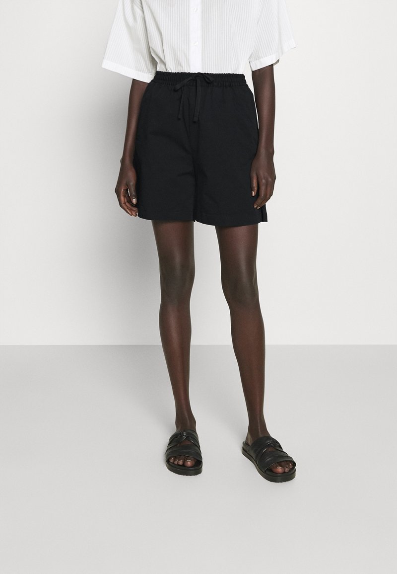 Person wearing black drawstring shorts, black slip-on sandals, and a white short-sleeve button-up shirt, standing on a plain light background.