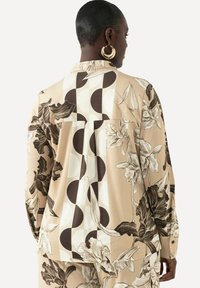 Blouse à manches longues en tissu beige avec des motifs floraux et géométriques en marron et crème. Coupe loose avec un détail de pli au dos.