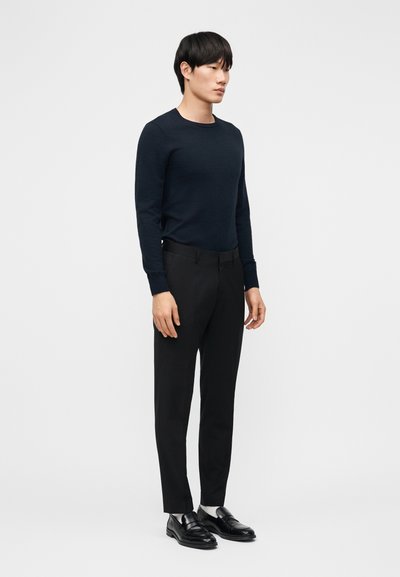 Jonge man staand, gekleed in een nauwsluitend zwart shirt met lange mouwen, zwarte broek, witte sokken en zwarte leren loafers, tegen een effen achtergrond.