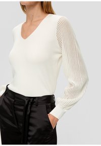 Pull blanc à col en V avec manches rayées transparentes texturées ; associé à un pantalon en satin noir avec taille nouée et poches latérales.