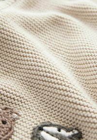 Beige stickad tyg med en texturerad mönster. Har broderade djurmotiv i brunt och grått i det nedre högra hörnet.