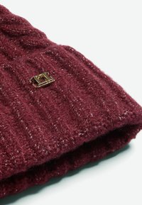 Căciulă tricotată de culoare bordó, cu o textură ribbed și un accent cu logo auriu în față. Materialul pare moale și călduros.