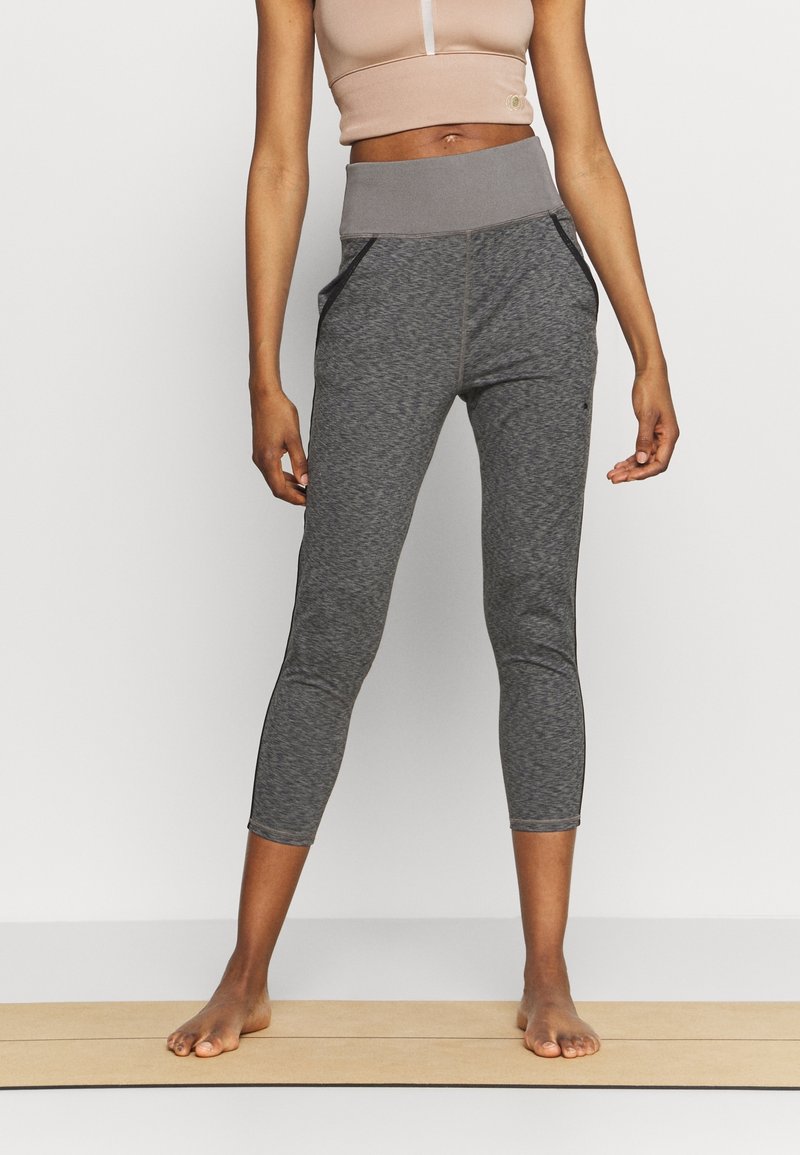 Mujer vestida con leggings cortos de cintura alta grises y una camiseta sin mangas corta beige, de pie descalza sobre una esterilla de yoga.