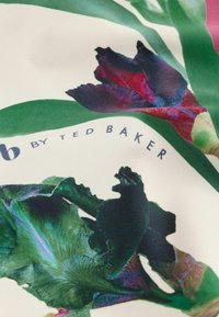 Zbliżenie tkaniny z fioletowo-zielonym nadrukiem kwiatowym i napisem "by Ted Baker" na białym tle.