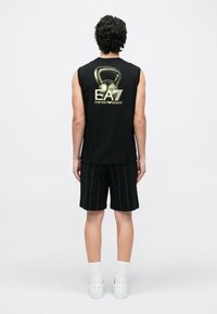 Άνδρας που φοράει μαύρο αμάνικο μπλουζάκι με γραφικό kettlebell και λογότυπο EA7 Emporio Armani, μαύρο σορτς, λευκές κάλτσες και λευκά αθλητικά παπούτσια, όρθιος με την πλάτη προς τα εμάς.