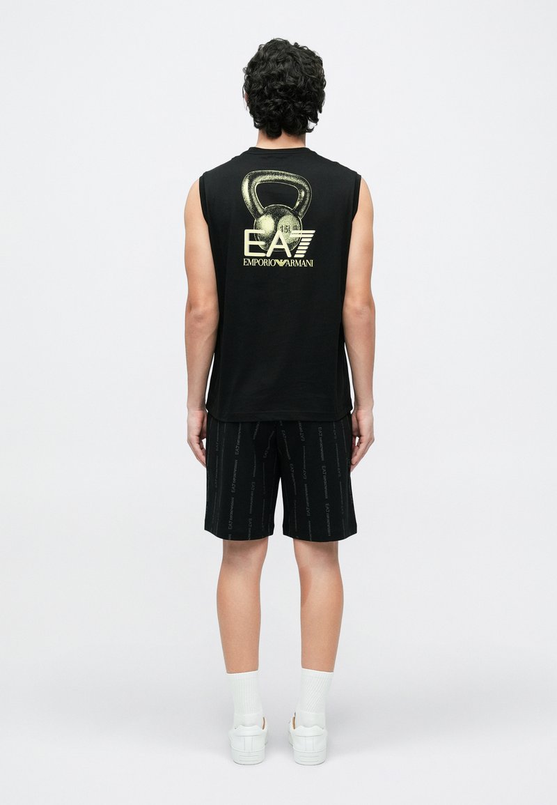 Άνδρας που φοράει μαύρο αμάνικο μπλουζάκι με γραφικό kettlebell και λογότυπο EA7 Emporio Armani, μαύρο σορτς, λευκές κάλτσες και λευκά αθλητικά παπούτσια, όρθιος με την πλάτη προς τα εμάς.