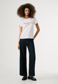T-shirt blanche à manches courtes avec un logo "Pepe Jeans London" rose et doré, associée à un pantalon large en denim foncé et des chaussures noires.