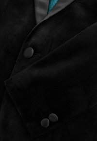 Gros plan sur une veste en velours noir, présentant une texture lisse, un col en satin et deux boutons ronds noirs sur le poignet.