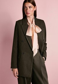 Mørkegrøn dobbeltradet blazer med sorte knapper, parret med et lys rosa silkebowtie og matchende bukser. Glat tekstur, skræddersyet pasform.