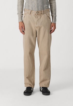 Osoba ubrana w beżowe spodnie o szerokich nogawkach, czarne trampki z białymi paskami oraz szary prążkowany top z długim rękawem, stojąca na białym tle.