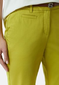 Felle limoengroene broek met een gladde textuur, voorzien van een vo Carpenterzak en riemlussen. Een bruine riem met een zilveren gesp accentueert de look.