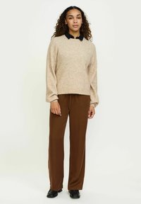 Svetlo bež pleten svetnik z dolgimi rokavi, v kombinaciji z rjavimi širokimi hlačami. Svetnik ima sproščen kroj in zaobljen ovratnik.