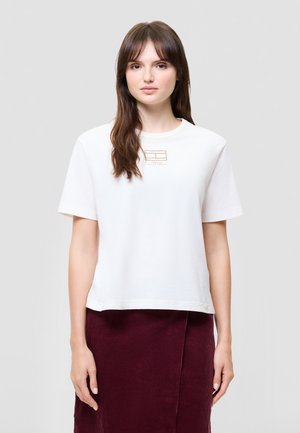 Une femme porte un T-shirt blanc à manches courtes orné d’un petit logo sur la poitrine et une jupe bordeaux, sur un fond gris uni.