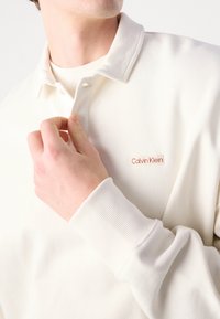 Biela polo košeľa s dlhým rukávom a ryhovaným golierom a manžetami. Na hrudi má malé červené logo "Calvin Klein". Vyrobená z mäkkej bavlny.