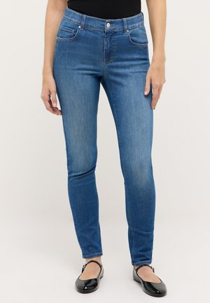 Donna che indossa jeans blu slim a vita media abbinati a scarpe nere piatte con cinturino alla caviglia su sfondo neutro.