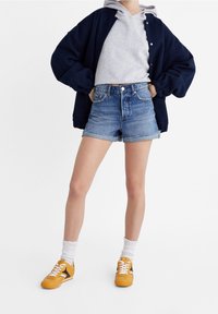 Pantalones cortos de mezclilla con dobladillo deshilachado, combinados con una sudadera gris y una chaqueta oversized azul marino. Zapatillas amarillas con detalles en negro y calcetines blancos.