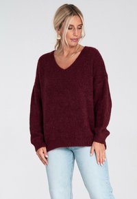 Kobieta z blond włosami, nosząca złote kolczyki i naszyjnik, ubrana w luźny, bordowy sweter w serek i jasnoniebieskie dżinsy, patrząca w dół.