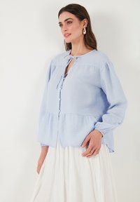 Blusa azzurra chiara realizzata in tessuto testurizzato, con maniche lunghe, chiusura frontale con bottoni e un nastro al collo. Dettaglio con orlo peplum.