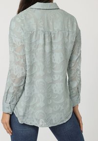 Blusa de color azul claro con un patrón floral texturizado, de mangas largas y con cuello. Presenta un dobladillo redondeado y detalles de botones en las mangas.