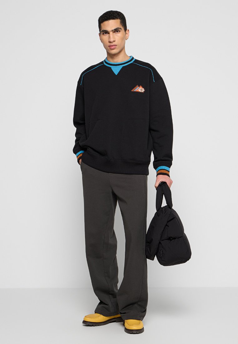 Czarna bluza z turkusowymi i pomarańczowymi akcentami, luźny krój, ściągacze przy rękawach i u dołu. Szare spodnie i żółte buty dopełniają stylizację, trzymając czarną torbę.