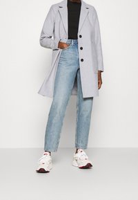 Grå uldfrakke med spids krave, sorte knapper, sidelommer, båret over en sort striktop, parret med lys blå højtaljede jeans og hvide sneakers.