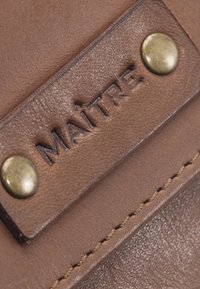 Accessorio in pelle marrone con etichetta embossée "MAÎTRE", hardware in ottone, cuciture a vista e superficie testurizzata.
