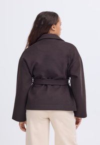 Donna con lunghi capelli scuri indossa una giacca marrone scuro con cintura e pantaloni beige chiaro, di spalle contro uno sfondo chiaro e uniforme.
