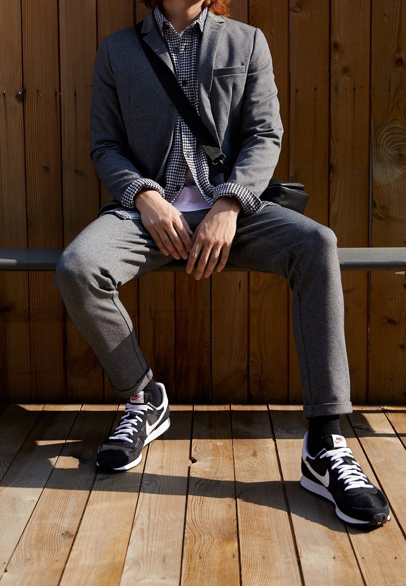 Personne assise sur un banc portant un costume gris, une chemise à motif pied-de-poule, des baskets Nike noires et blanches, avec un sac bandoulière noir contre un mur en bois.