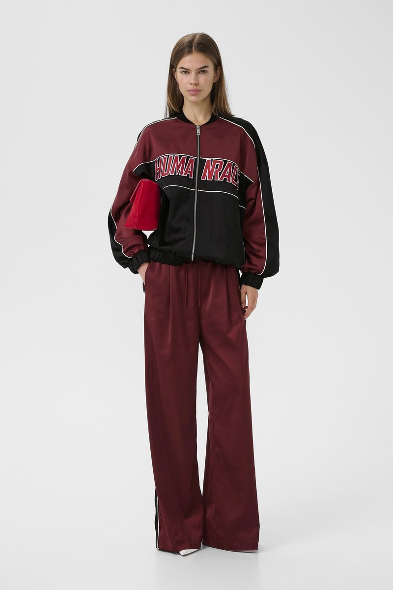 Woman wearing a burgundy and black zip-up jacket with "HUMANRACE" text, matching wide-leg pants, holding a red clutch bag.