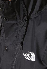 Närbild av svart utomhusjacka med justerbar huva och dragsko samt "The North Face"-logotyp p å bröstet.