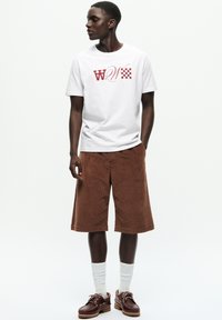 Tricou alb cu imprimeu grafic roșu, pantaloni scurți din bumbac piersici de culoare cafenie, șosete albe și saboți maro cu talpă din lemn. Modelul stă drept.