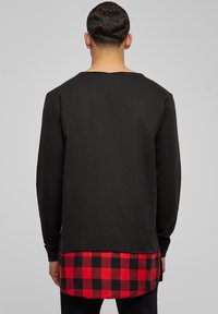 Chemise à manches longues noire avec un ourlet en tissu à carreaux rouges et noirs, présentant des détails de couture visibles. Tissu doux et coupe décontractée.