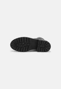 Semelle de botte noire avec des sculptures profondes présentant des motifs rectangulaires et en forme de croix sur un fond gris clair.