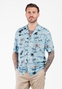 Uomo con barba e tatuaggi che indossa una camicia a maniche corte azzurro chiaro con motivi di palme e velieri e pantaloni beige, con le mani in tasca.