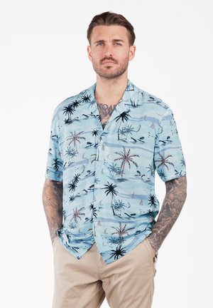 Beroepsbeeld met een baard, tattoos, en een lichtblauw kortemouwen shirt met palmbomen en zeilbootpatronen, gecombineerd met beige broek, handen in de zakken.
