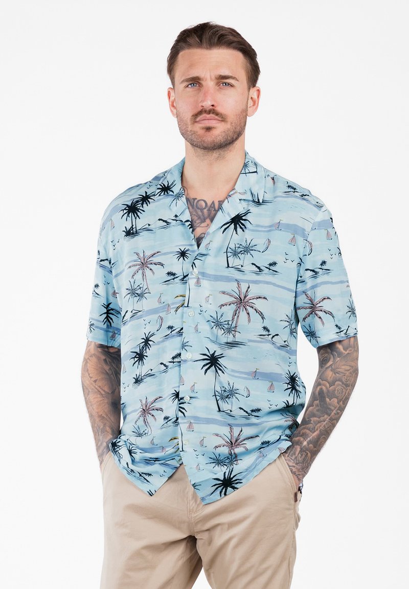 Uomo con barba e tatuaggi che indossa una camicia a maniche corte azzurro chiaro con motivi di palme e velieri e pantaloni beige, con le mani in tasca.