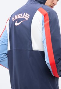 Homme portant une veste bleu marine, bleu clair, blanche et rouge avec "England" et un logo Nike au dos, légèrement tourné sur le côté.