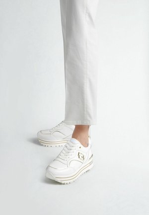 Witte plateau sneakers met gouden accenten, gedragen met witte cropped broek op een neutrale achtergrond.